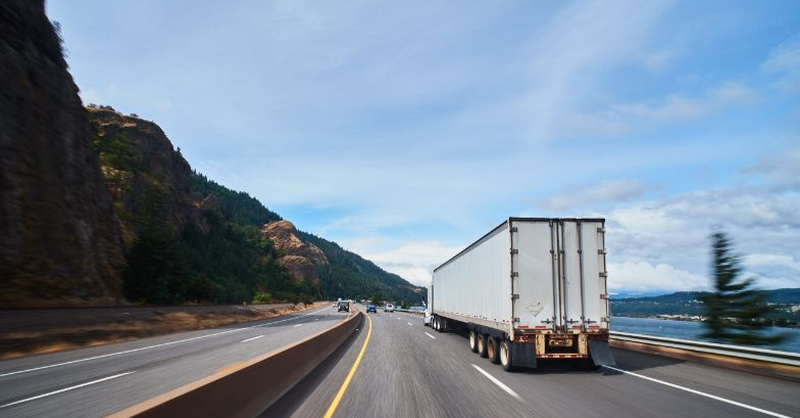 Semi‑truck driving on a winding highway through a mountainous, scenic landscape.
