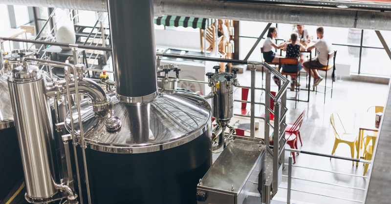 **Alt text:**   Large stainless-steel brewing equipment inside a modern brewery overlooks a seating area where several people are gathered at a table on an upper level.
