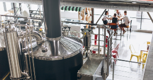 **Alt text:**   Large stainless-steel brewing equipment inside a modern brewery overlooks a seating area where several people are gathered at a table on an upper level.