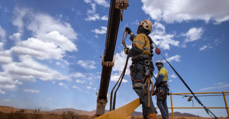 Workers wearing safety harnesses use a self‑retracting lifeline while working at height on a steel structure.