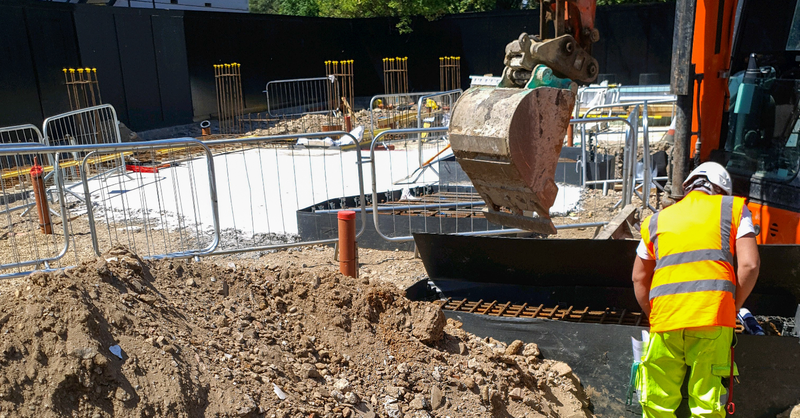 Construction worker stands beside an excavator digging a trench at a construction site.