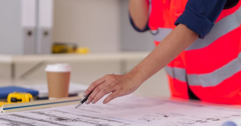 A design professional reviewing construction drawings on a desk in a modern office, representing ongoing project coordination, documentation review and post‑pandemic risk management in construction.