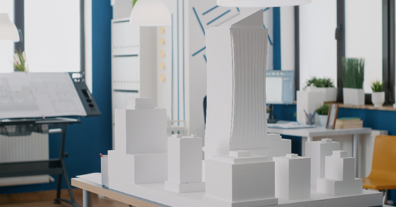 An architectural scale model of modern buildings sits on a table in a bright design office, surrounded by drafting tools and plans, representing architectural planning, design development and institutional continuity in a professional workspace.