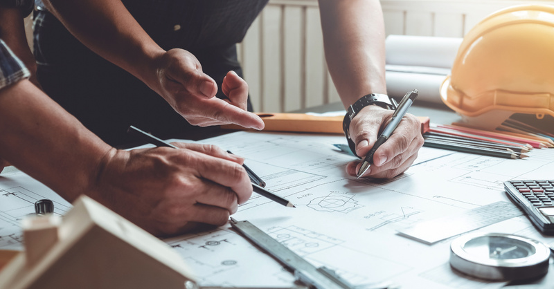 Two people review architectural plans on a table with drafting tools and a hard hat.