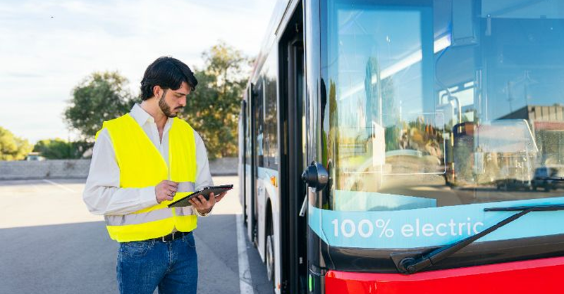 A safety‑vested worker stands beside an electric bus while reviewing information on a tablet