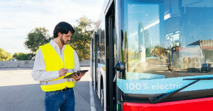 A safety‑vested worker stands beside an electric bus while reviewing information on a tablet.