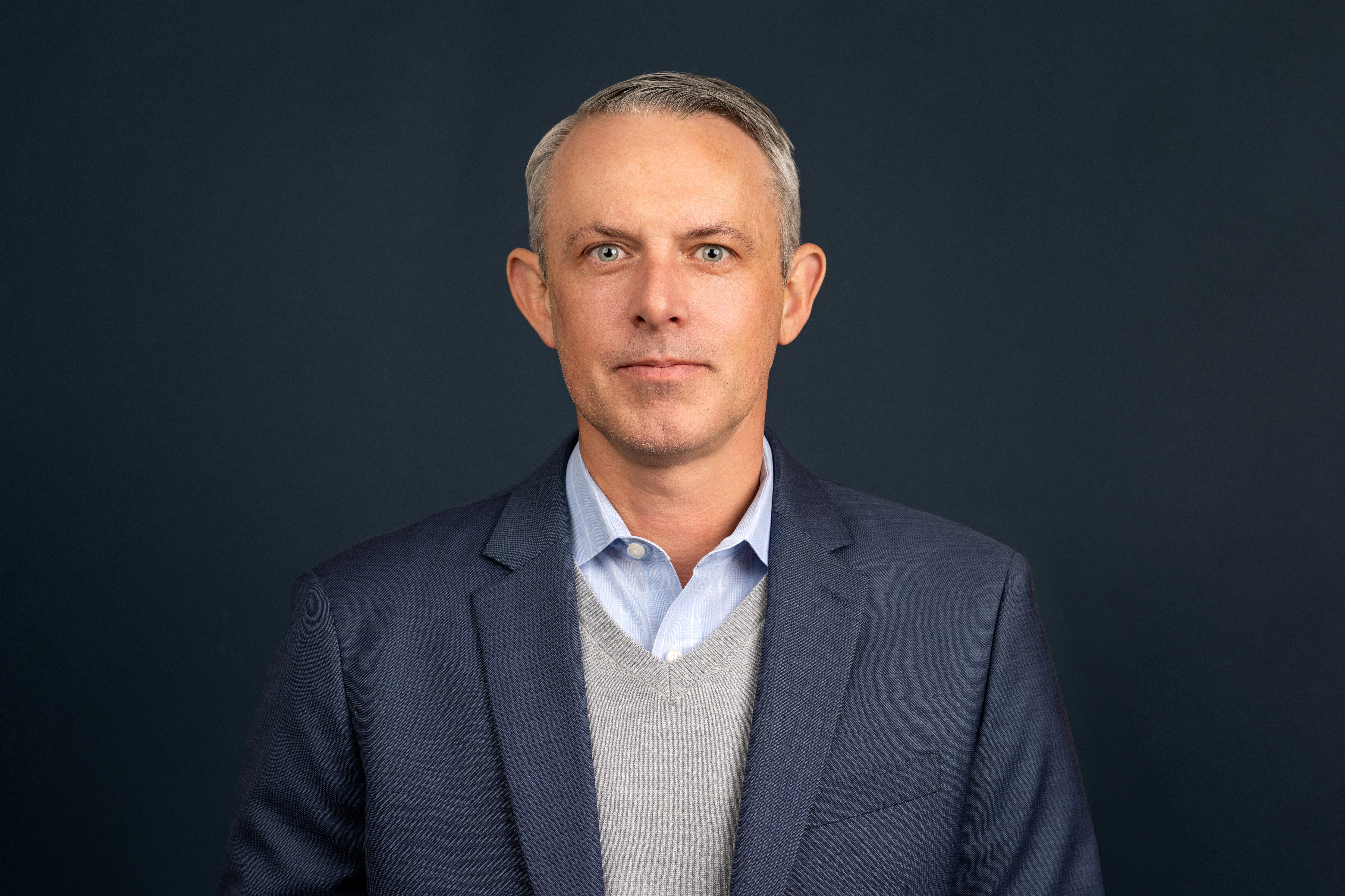 Professional studio portrait of a person wearing a navy blazer over a light gray sweater and light blue collared shirt, standing against a dark, neutral background.