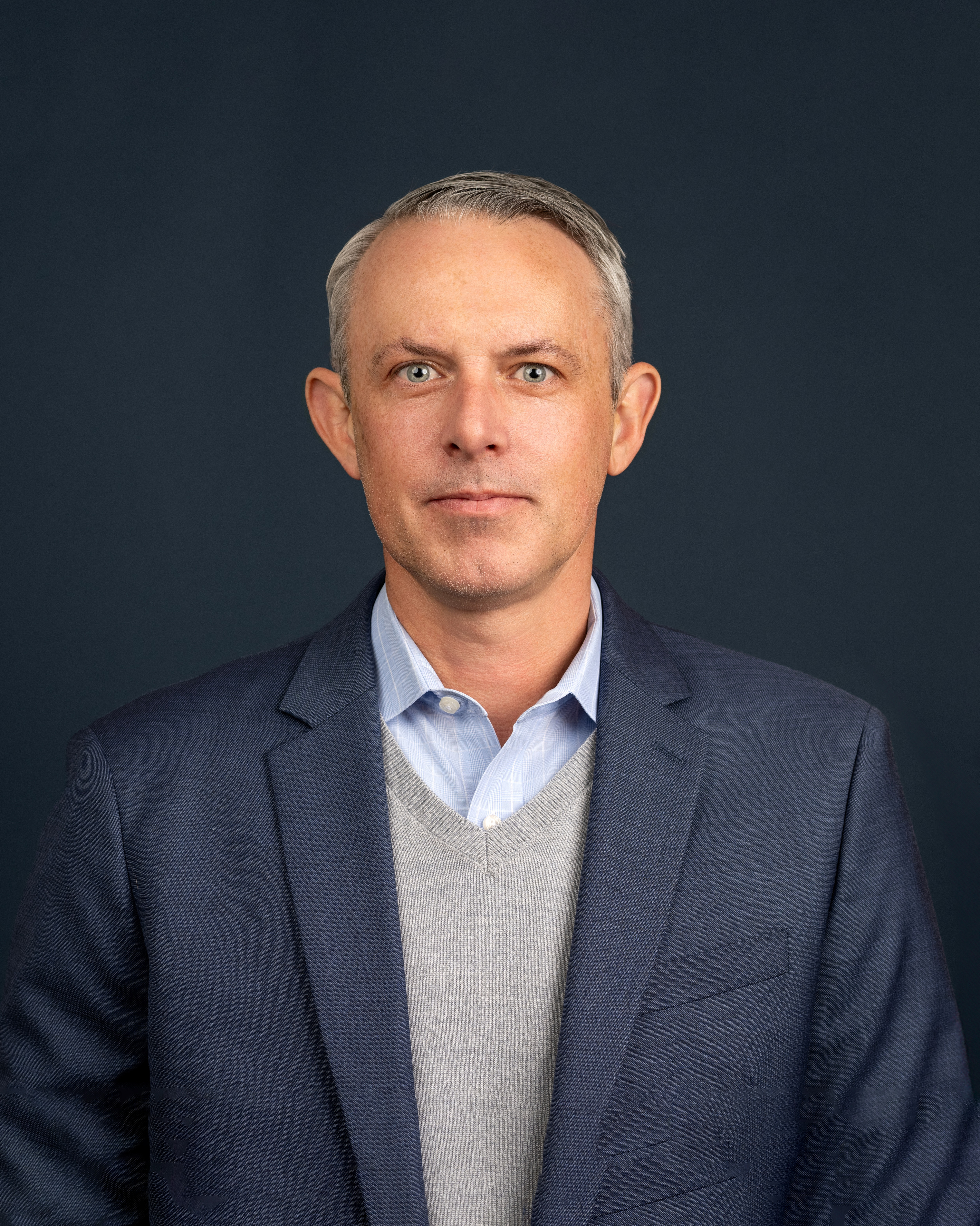 Professional studio portrait of a person wearing a navy blazer over a light gray sweater and light blue collared shirt, standing against a dark, neutral background.