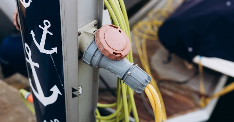 A marina power pedestal with electrical connectors and cables on a dock beside a moored boat, highlighting shore power equipment and electrical safety at a marina.
