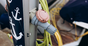 A marina power pedestal with electrical connectors and cables on a dock beside a moored boat, highlighting shore power equipment and electrical safety at a marina.