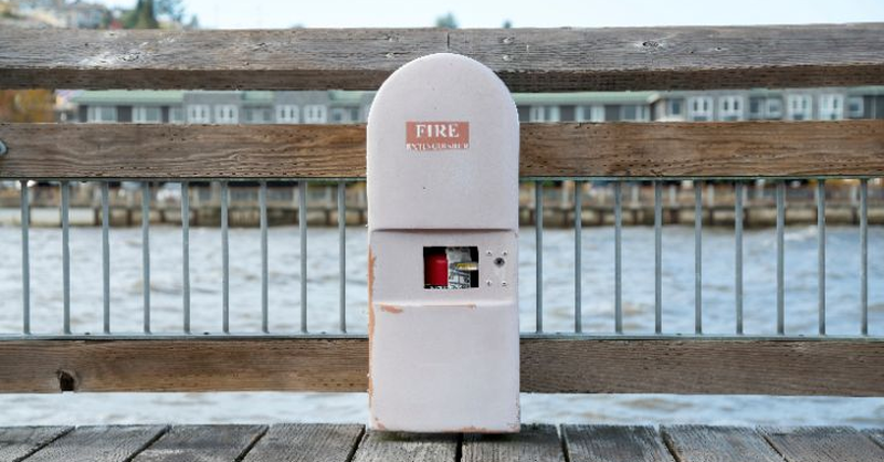 Fire extinguisher inside a labeled cabinet on a marina dock, with water and waterfront buildings in the background, representing marina fire safety, emergency preparedness, and fire prevention measures.