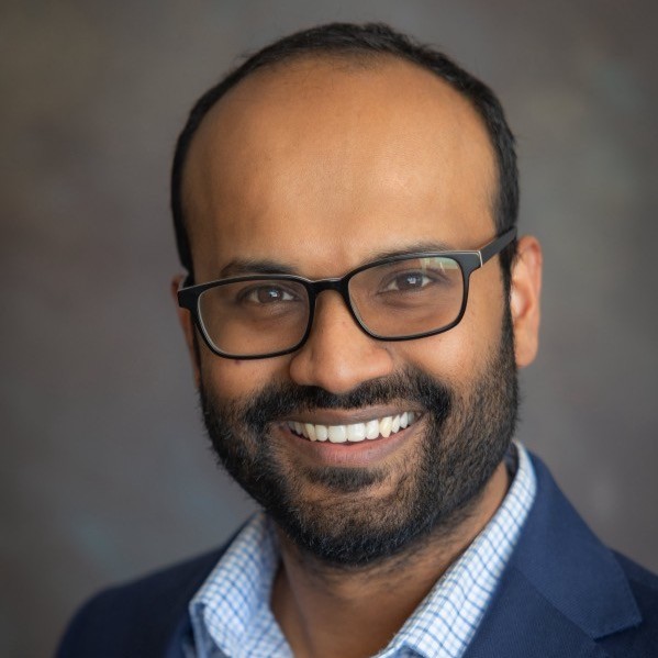 Professional headshot of a person wearing black rectangular glasses and a dark blazer over a light checkered shirt, facing the camera with a smile, against a softly blurred gray background.