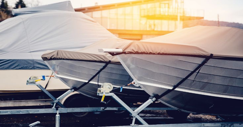 Two boats covered with protective tarps sit on trailers in front of a building, illuminated by warm sunlight.