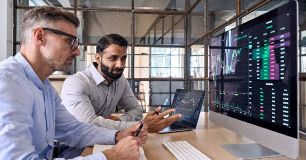 Two colleagues sit at a desk reviewing data visualizations displayed on large computer monitors. Charts and graphs on the screens show financial or analytical trends as they discuss insights together.